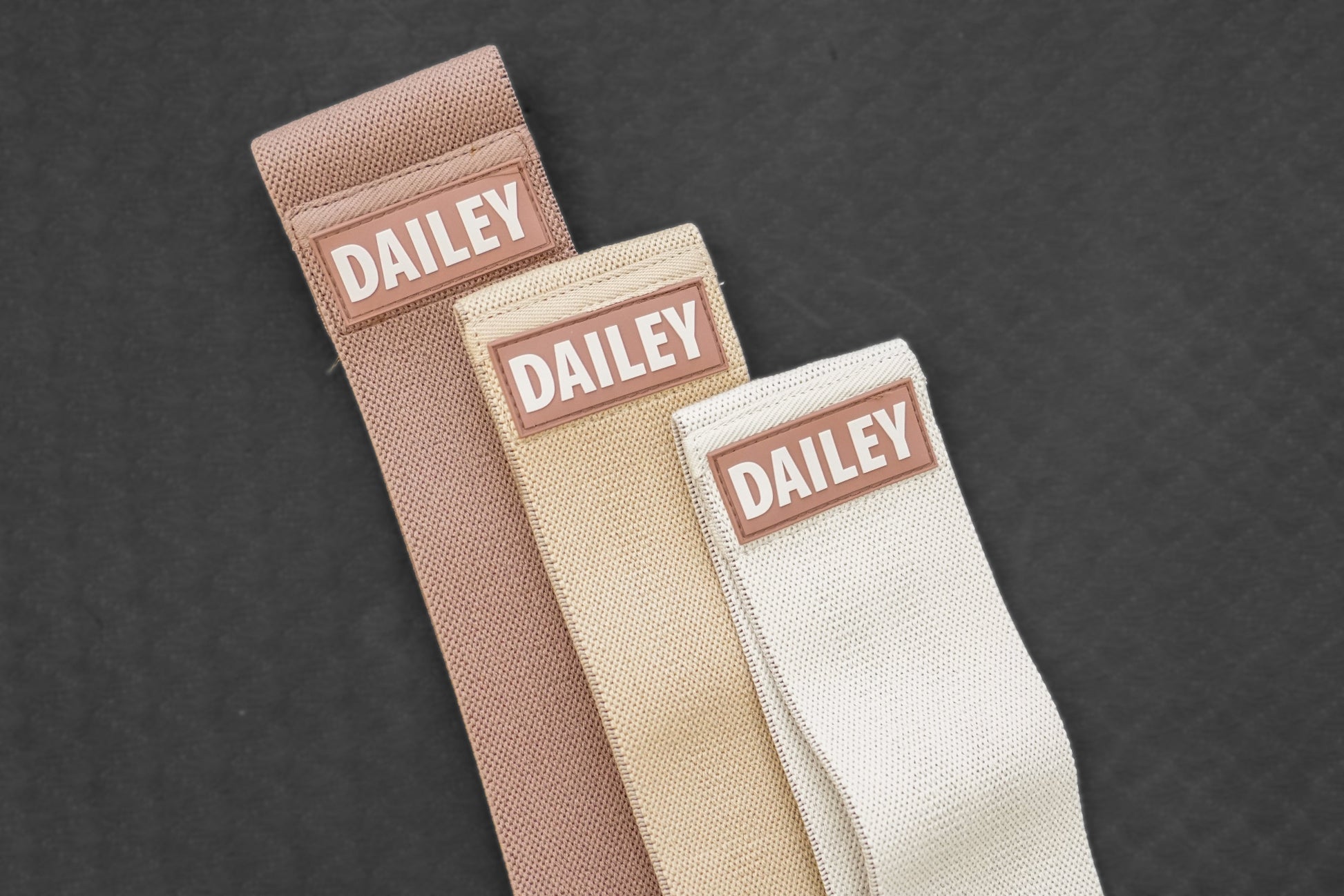 Three woven fabric straps in dusty pink, beige, and off-white, each featuring a rectangular brown patch with the word 'DAILEY' in white, arranged diagonally on a dark textured background.